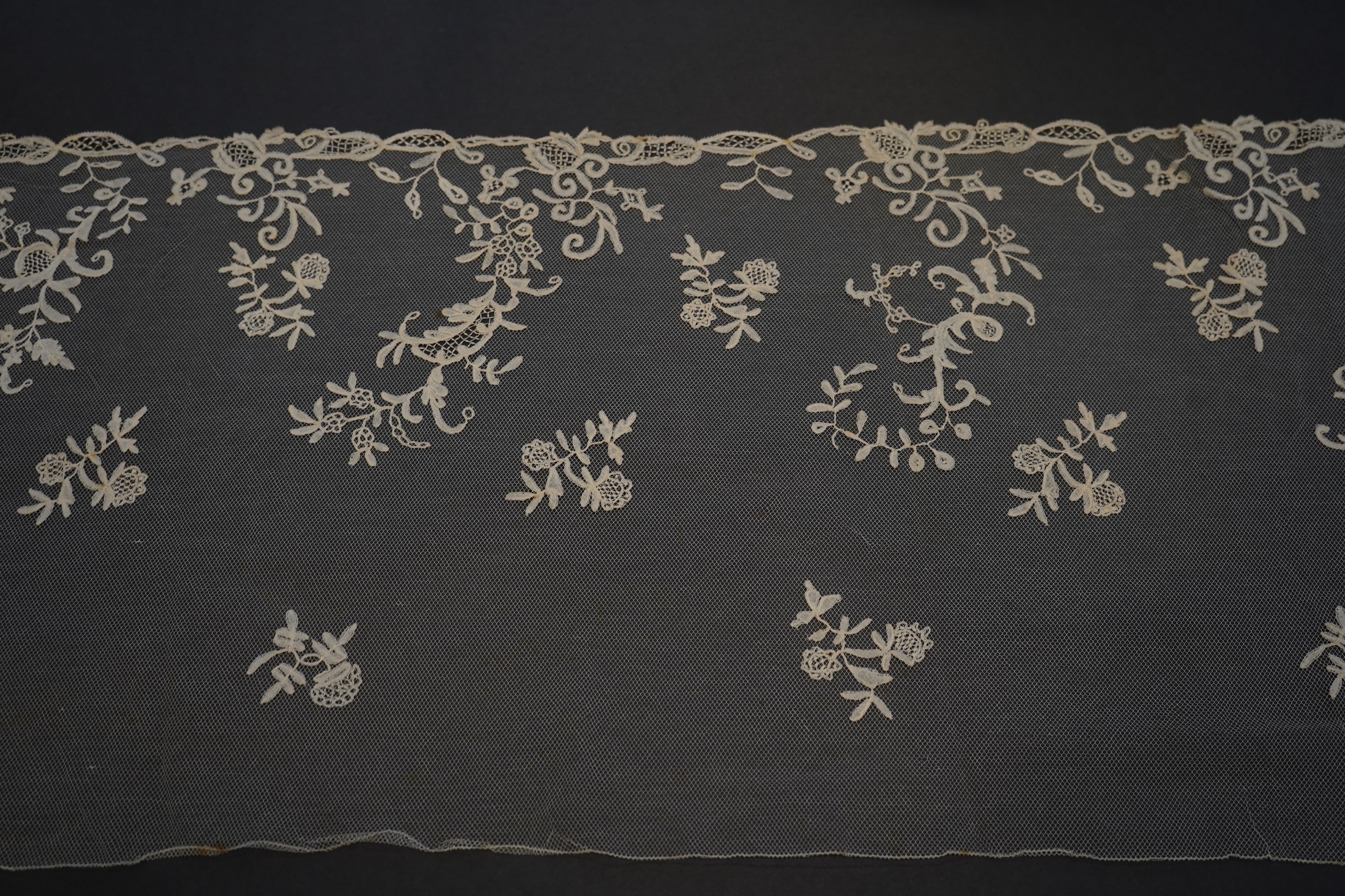 A 19th century five yard flounce of Brussels bobbin appliqué lace applied onto fine machine net, worked in a trailing floral design with an ornate border and sprig motifs, 457.5 cm long x 36cm deep. Condition - possibly